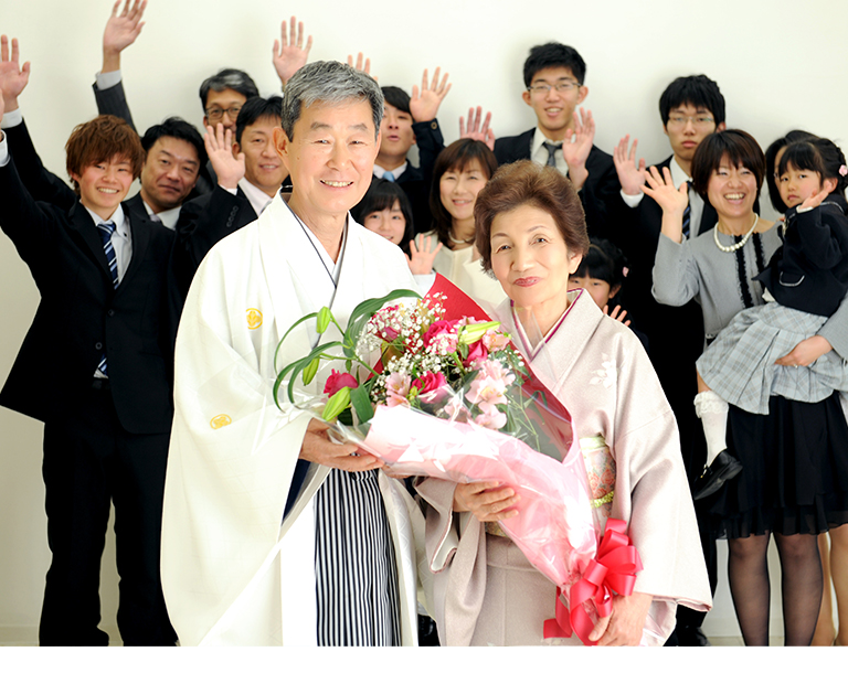 花束を持つ着物姿の夫婦と拍手するご親族の写真